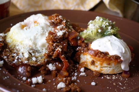 Closeup on masa harina biscuits with spicy chorizo topping. Two halves of a biscuit are each covered in a generous scoop of chorizo topping that spills over onto the plate; the left half is topped with a fried egg, and the right with sour cream and guacamole; crumbled cotija cheese is sprinkled over the whole plate.