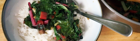 A bowl of rice topped with "simple Swiss chard with garlic and crystallized ginger," sits on a wooden countertop. Next to it sits a stainless steel pan filled with more chard, the bright red and yellow stems punctuated the emerald-green leaves.