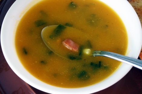 Closeup on a bowl of butternut squash soup with ham an scallions. The soup is a deep gold color, studded with dark green scallions, half submerged. A spoon rest in the soup, its bowl partially lifted out, supporting a nubbin of pink ham and a round of pale-green scallion.