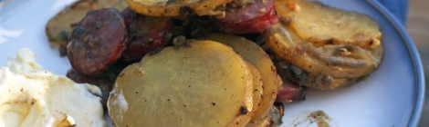 Closeup on a plate of fried potatoes, pale yellow discs of potato studded with brick-red rounds of sausage and soft shreds of meltingly soft jalapeño and onion. In the foreground, blurry, a bite potato and sausage speared on a fork, touched with mayonnaise from a thick dollop in the corner of the frame.