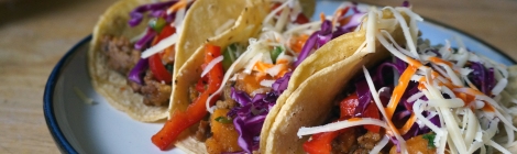 Sweet potato tacos with marinated peppers and cabbage, three of them, nestled together on a plate, garnished with white shredded cheese and bright orange hot sauce.