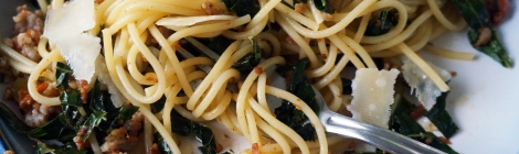 A fork lies amidst a tangle of spaghetti, tines completely hidden. Ribbons of kale, small nubbins of sausage, and shards of Parmesan cheese punctuate the plate.